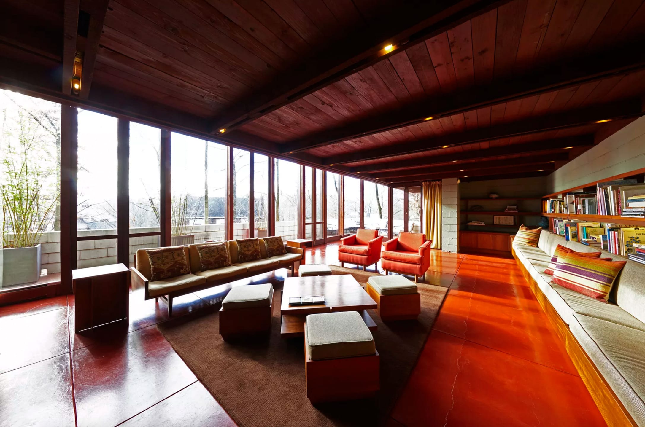 Spacious living room with wooden ceiling, large windows, orange floor, modern furniture, and built-in bookshelves.