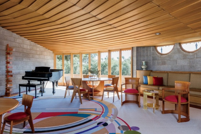 Spacious living room with round table, wooden chairs, colorful rug, grand piano, and large windows with wooded view.
