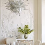 Modern dining room with intricate wall art, large floral chandelier, glass table, and decorative plants.