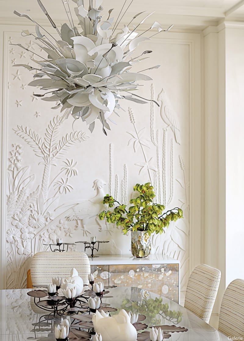Modern dining room with intricate wall art, large floral chandelier, glass table, and decorative plants.