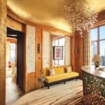 Luxurious room with golden walls, a yellow sofa, abstract rug, chandelier, and large windows offering a city view.