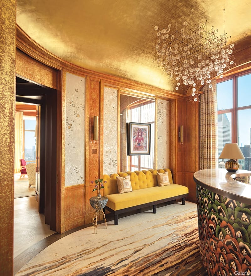 Luxurious room with golden walls, a yellow sofa, abstract rug, chandelier, and large windows offering a city view.