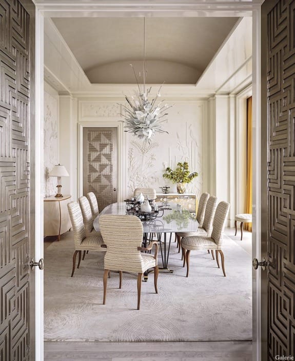 Elegant dining room with a glass table, patterned chairs, decorative chandelier, and textured walls.