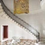 Elegant spiral staircase with black railing, geometric chandelier, abstract wall art, and modern white sculptural chair.