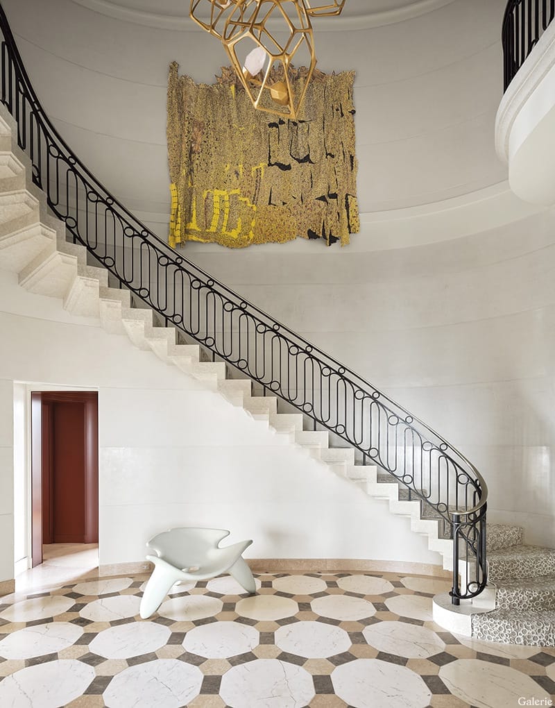 Elegant spiral staircase with black railing, geometric chandelier, abstract wall art, and modern white sculptural chair.