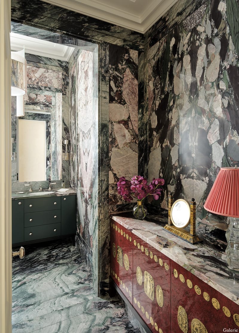 Luxurious bathroom with intricate marble walls, elegant furnishings, red dresser, and decorative lighting.