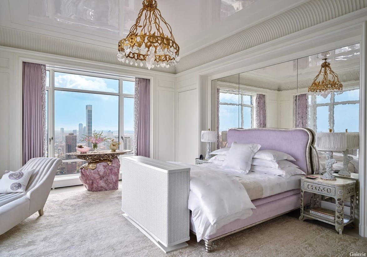 Luxurious bedroom with elegant decor, large windows with city view, plush bed, mirrored wall, and vintage chandeliers.
