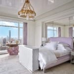 Luxurious bedroom with elegant decor, large windows with city view, plush bed, mirrored wall, and vintage chandeliers.
