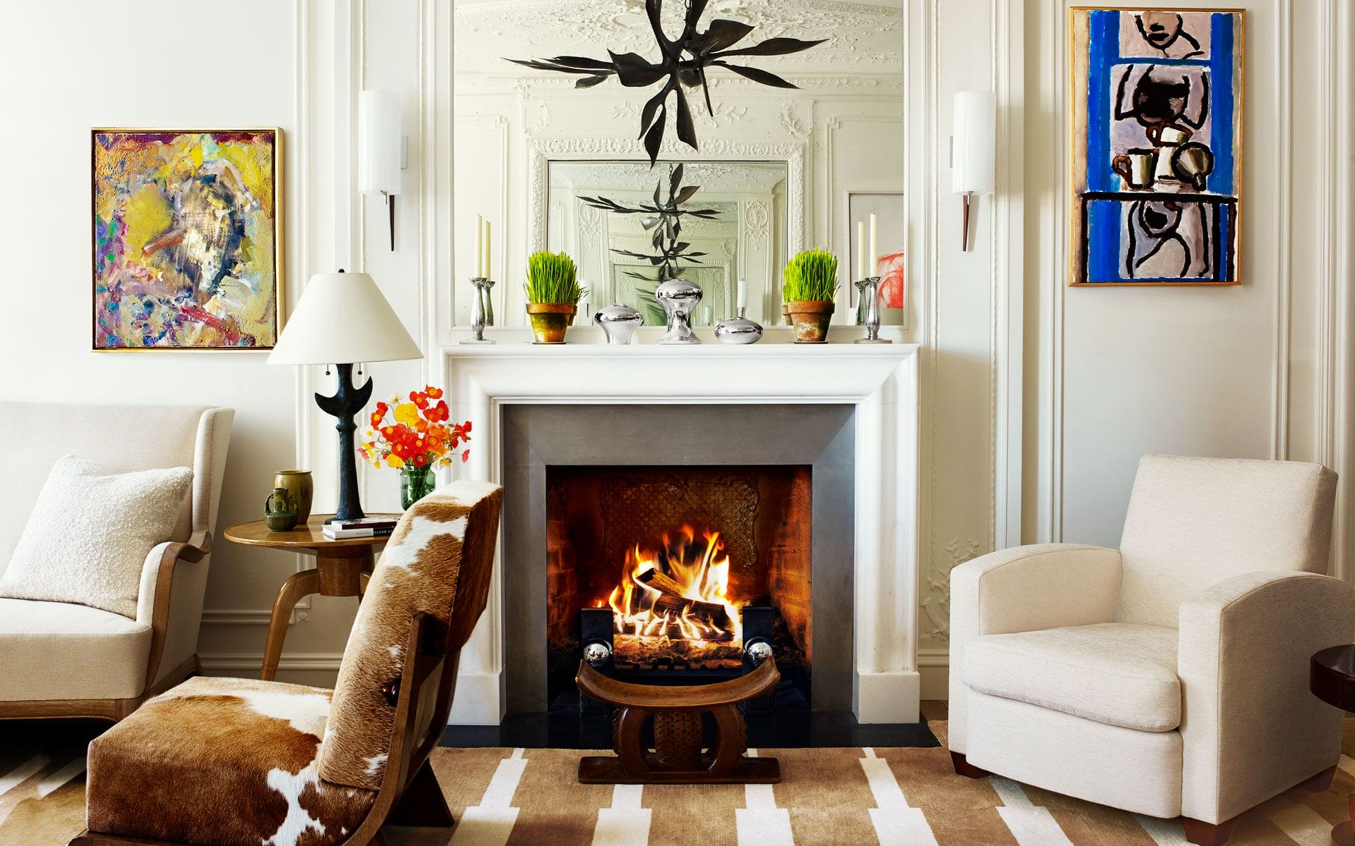 Cozy living room with a lit fireplace, modern art, white armchairs, and plants on the mantel.