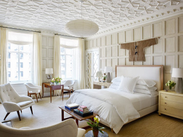 Elegant bedroom with white bedding, textured ceiling, armchairs, wooden furniture, large windows, and decorative wall hanging.