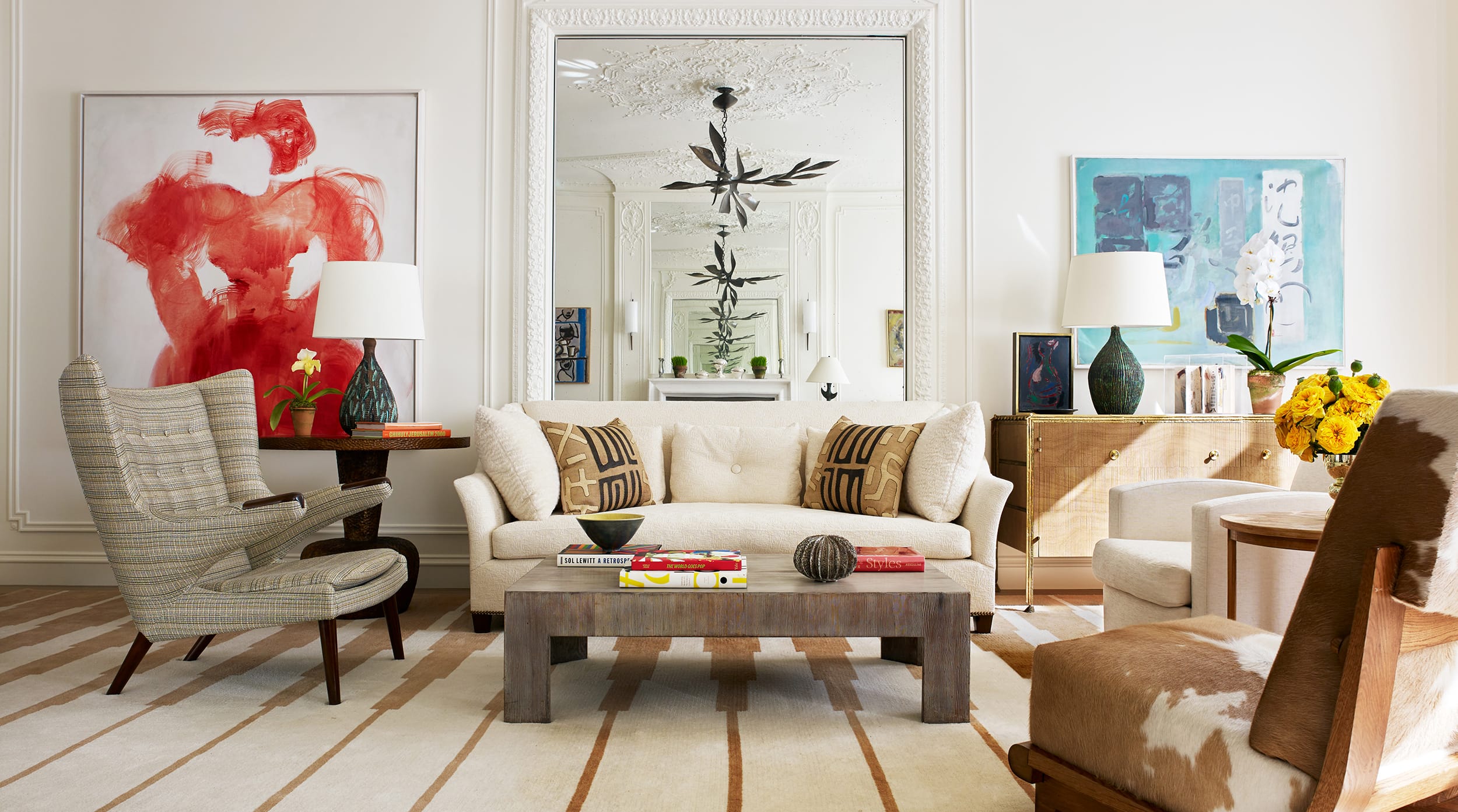 Elegant living room with cream sofa, striped rug, modern art, and decorative mirror, flanked by chairs and lamps.