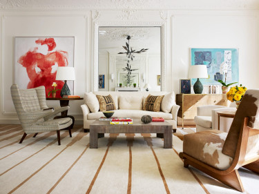 Elegant living room with beige sofas, modern art on walls, large mirror, and cozy chairs on a striped carpet.