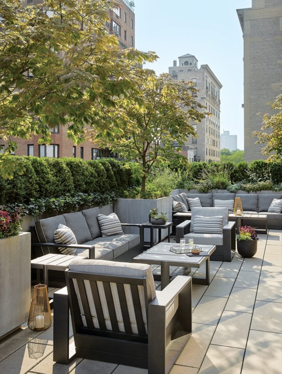 Rooftop patio with cushioned seating, lush greenery, and cityscape view on a sunny day.