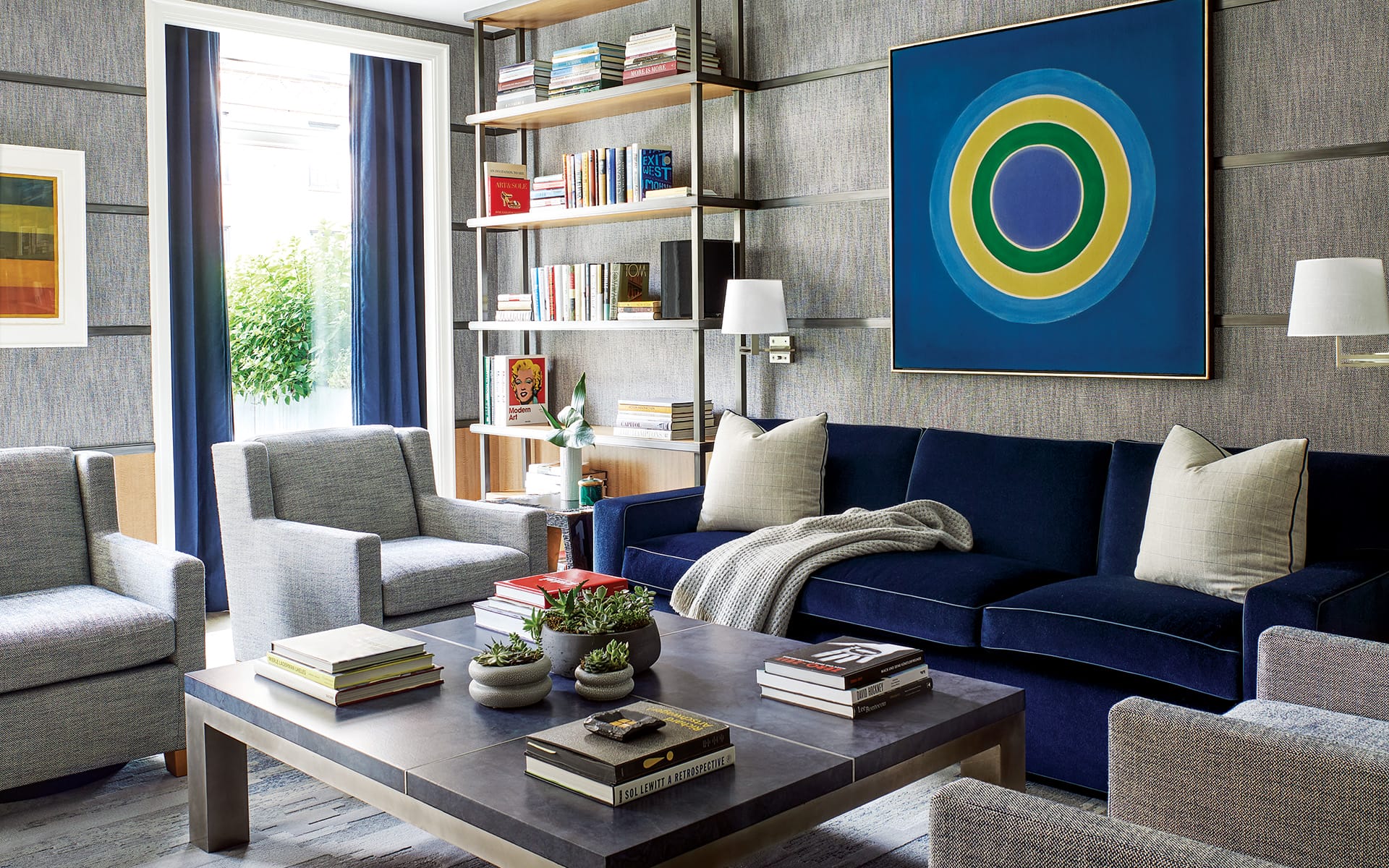 Modern living room with blue sofa, gray chairs, abstract wall art, books on wooden shelves, and a coffee table with decor.