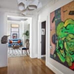 Modern hallway with wooden floors, vibrant abstract art on walls, and a view into a cozy living room with blue sofa and large windows.