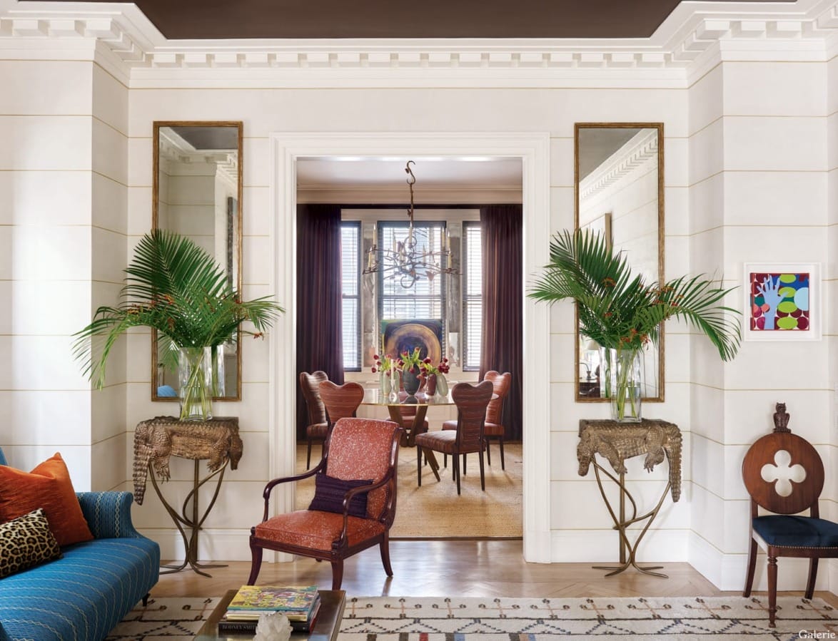 Elegant living room with vibrant furniture, tall plants, and mirrored accents leading to a dining area with a chandelier.