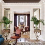 Elegant living room with vibrant furniture, tall plants, and mirrored accents leading to a dining area with a chandelier.