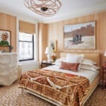 Bedroom with orange patterned wallpaper, modern art, and a geometric ceiling light fixture above a neatly made bed.