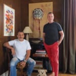 Two men sitting and standing in a room with artwork on walls, a small dog on the carpet, and books stacked between them.