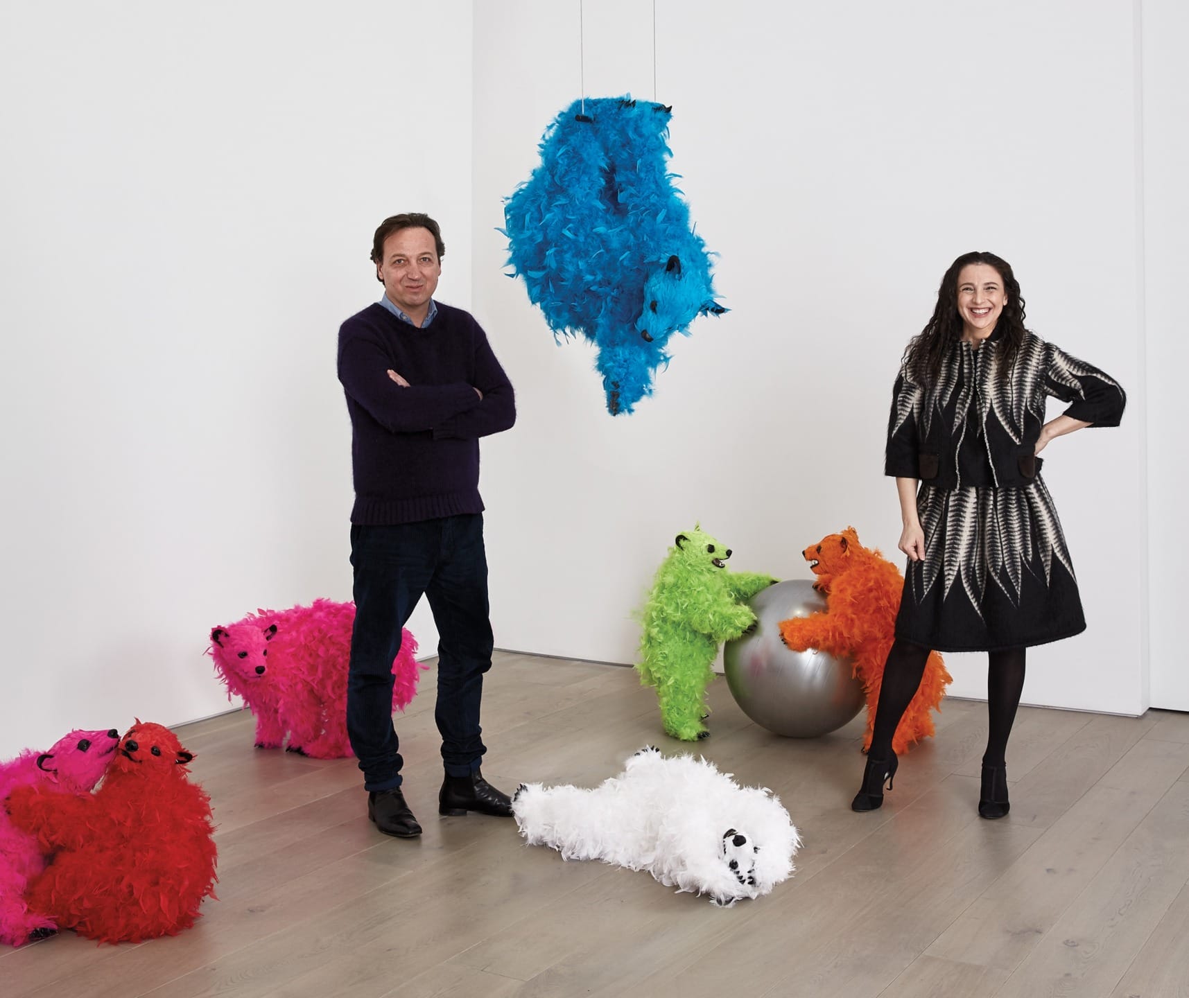 Two people standing in an art gallery with colorful, fluffy animal sculptures on the floor and one hanging.