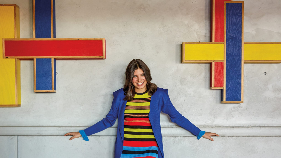 Person in colorful striped dress and blue coat standing between two abstract wall art pieces with red, blue, yellow frames.