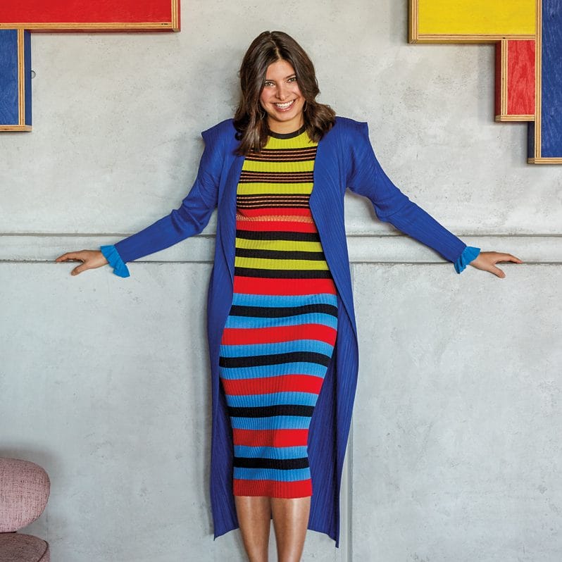 Woman in a colorful striped dress and blue coat stands smiling against a modern abstract background.