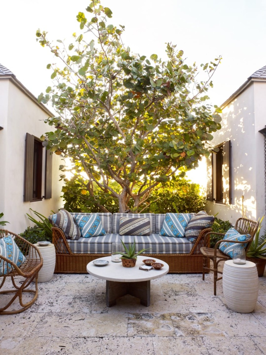 Cozy outdoor patio with striped sofa, decorative pillows, wicker chairs, coffee table, and lush greenery.