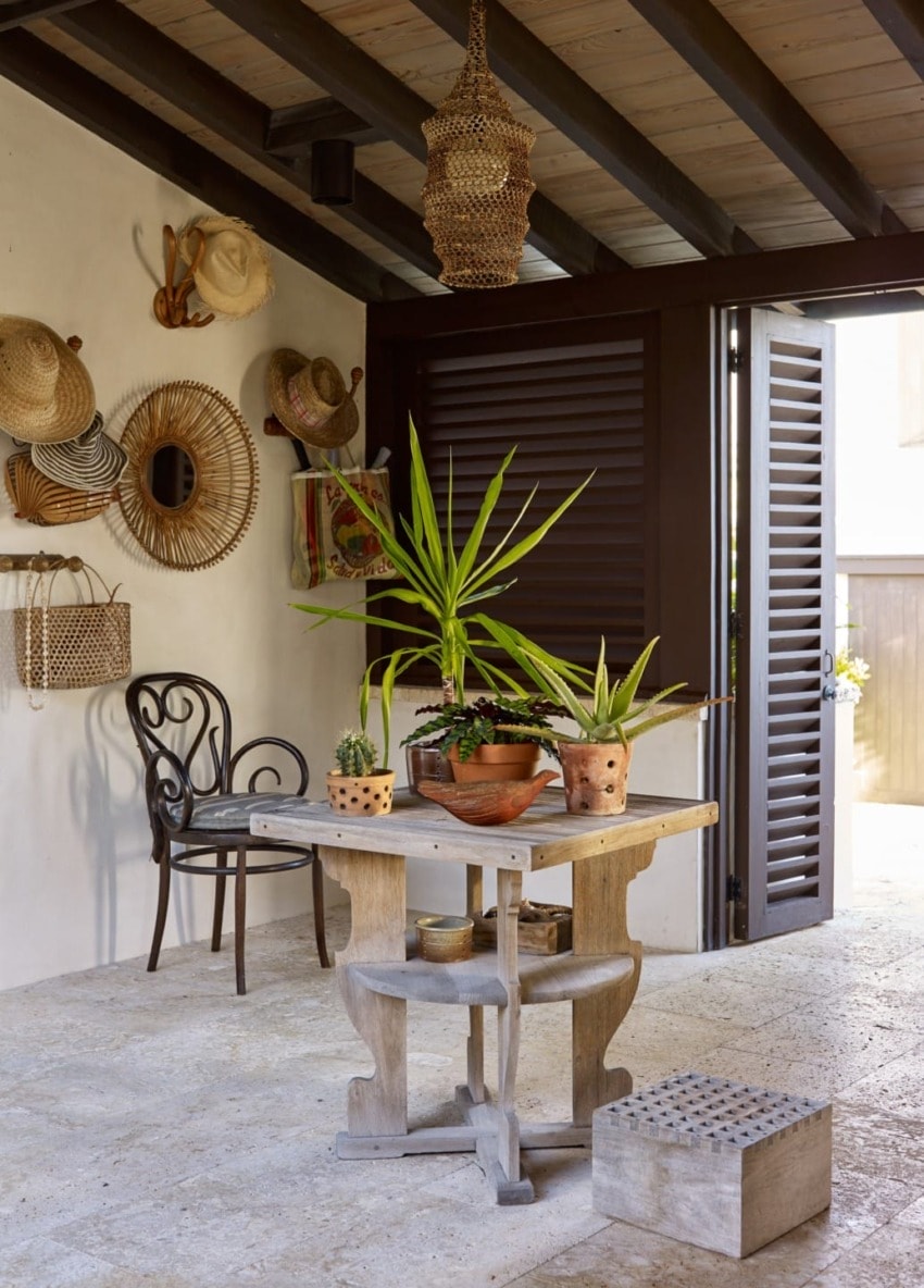 Cozy patio with a wooden table, plants, wicker decor, and hanging hats under a warm, rustic ceiling with wooden beams.