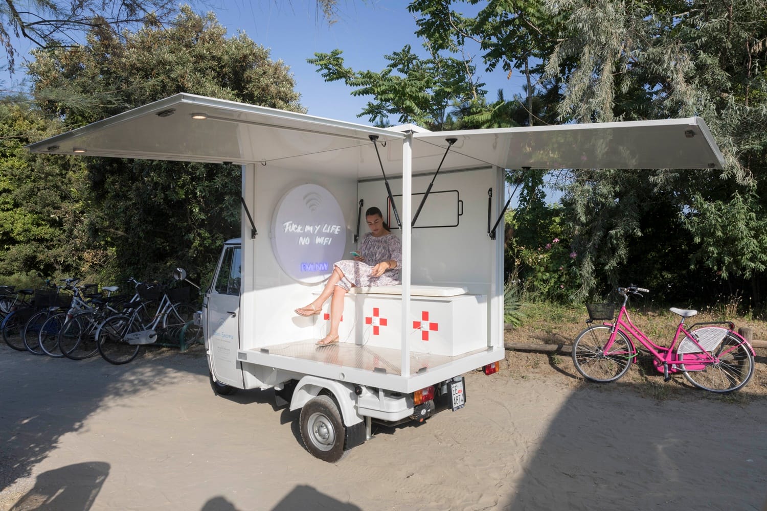 Small truck with open sides displaying a cozy interior space with a woman sitting inside and bicycles parked nearby.