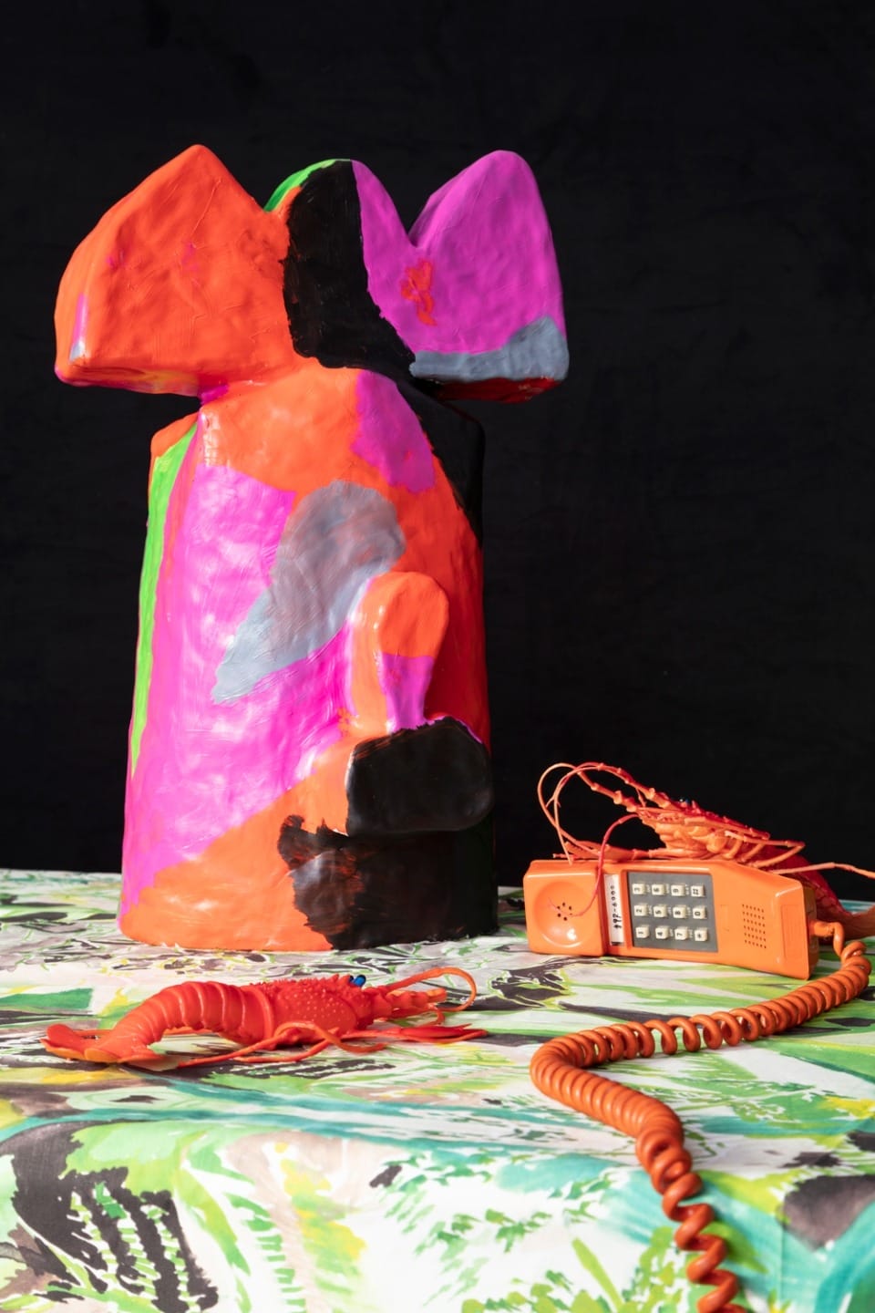 Colorful abstract sculpture on a patterned tablecloth with an orange vintage rotary phone and toy lobster.