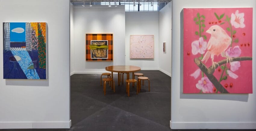 Art gallery with colorful paintings on walls and a round wooden table with stools in the center of the room.