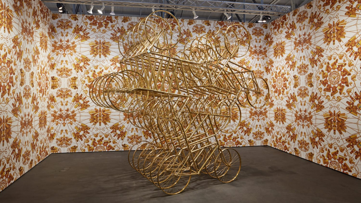 Abstract sculpture made of interconnected golden bicycle frames in a patterned exhibition space.