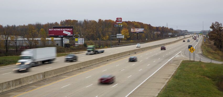 Cars and trucks driving on a highway surrounded by trees in autumn with billboards in the background.
