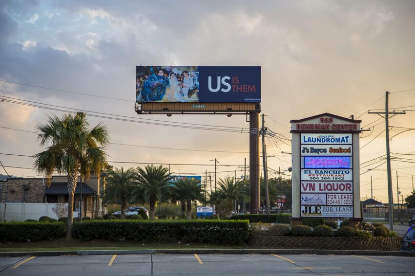 Billboard with "US IS THEM" message above a shopping center with palm trees and various store signs in the foreground.