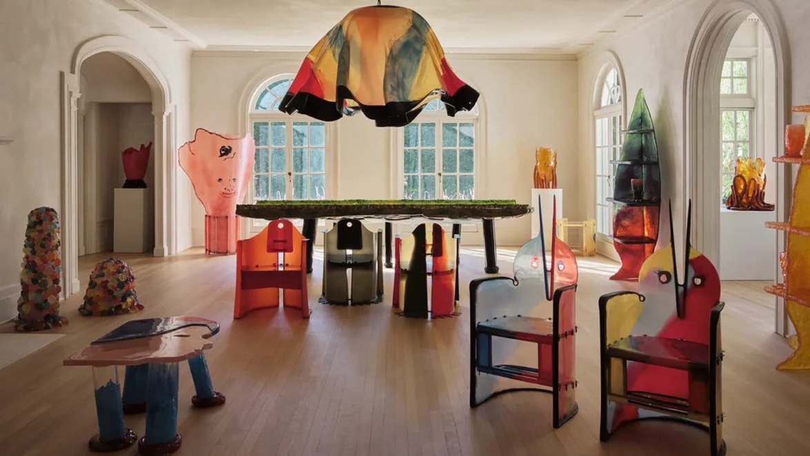 Colorful modern art exhibition in a bright room with unique chairs, a large table, abstract sculptures, and natural light.