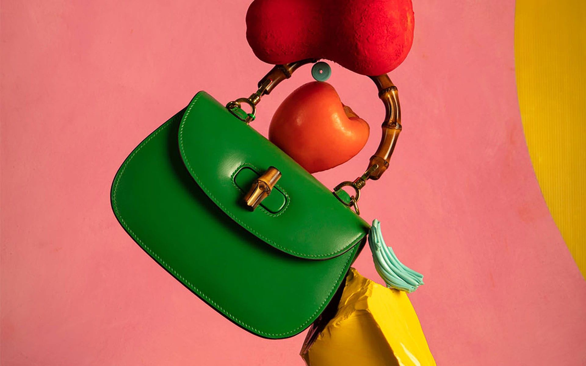 Green handbag with a bamboo handle, adorned by colorful abstract shapes against a vibrant pink background.