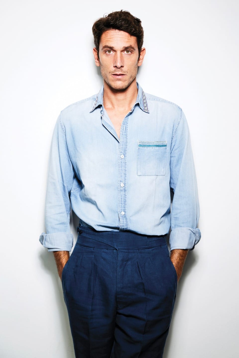 Man in a light blue denim shirt stands against a plain white background with hands in pockets, looking straight ahead.
