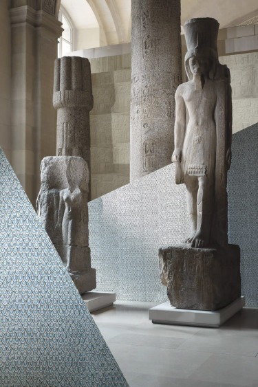 Ancient stone statues displayed in a museum setting with intricate wall patterns in the background.