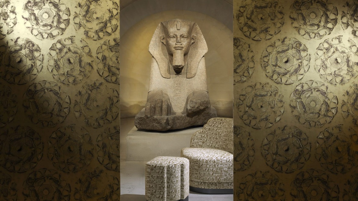Ancient Egyptian sphinx statue surrounded by decorative patterned walls and furniture in a museum setting.
