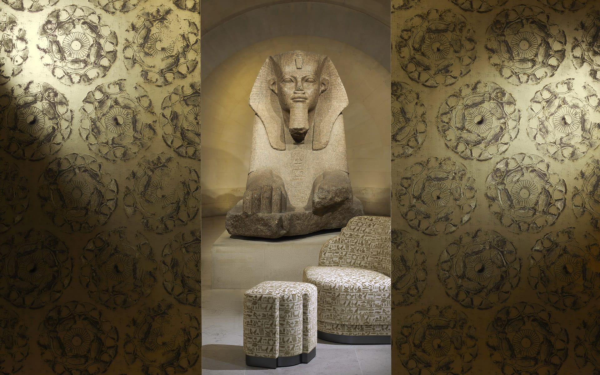 Ancient Egyptian sphinx statue surrounded by decorative patterned walls and furniture in a museum setting.