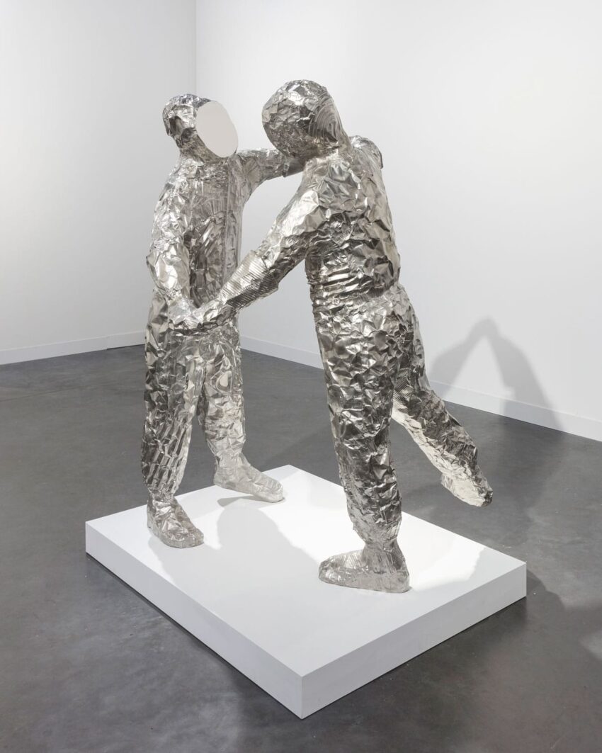Abstract silver sculpture of two faceless figures in a pose, displayed on a white platform in a gallery setting.