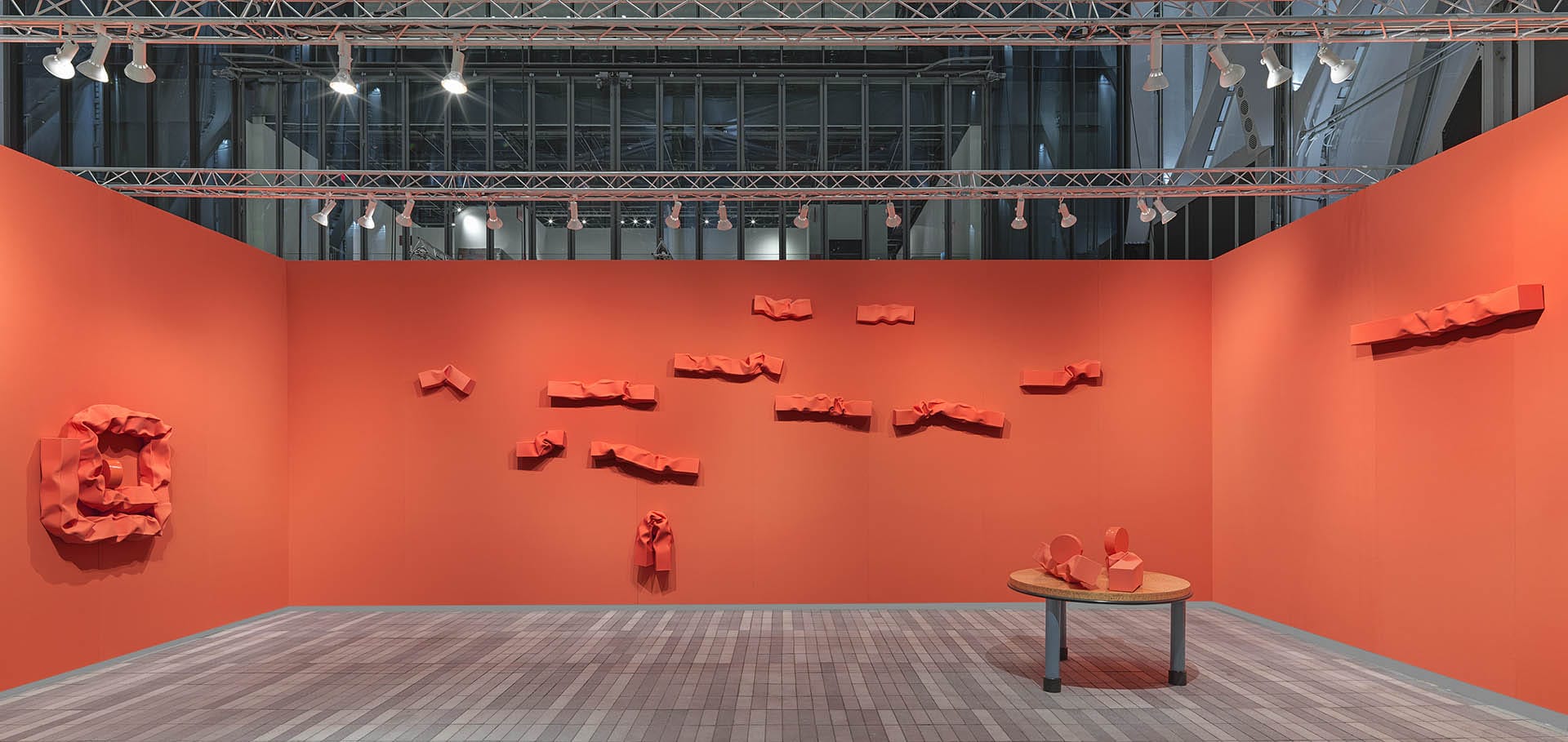 Gallery with orange walls displaying abstract red sculptures mounted and on a table, under bright overhead lighting.