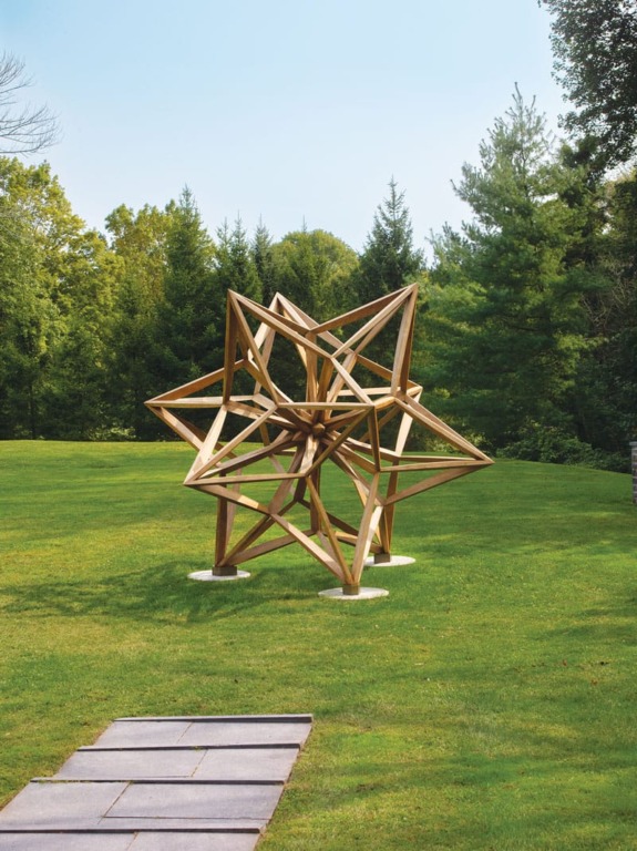 Wooden geometric sculpture on a grassy field with surrounding trees under a clear blue sky