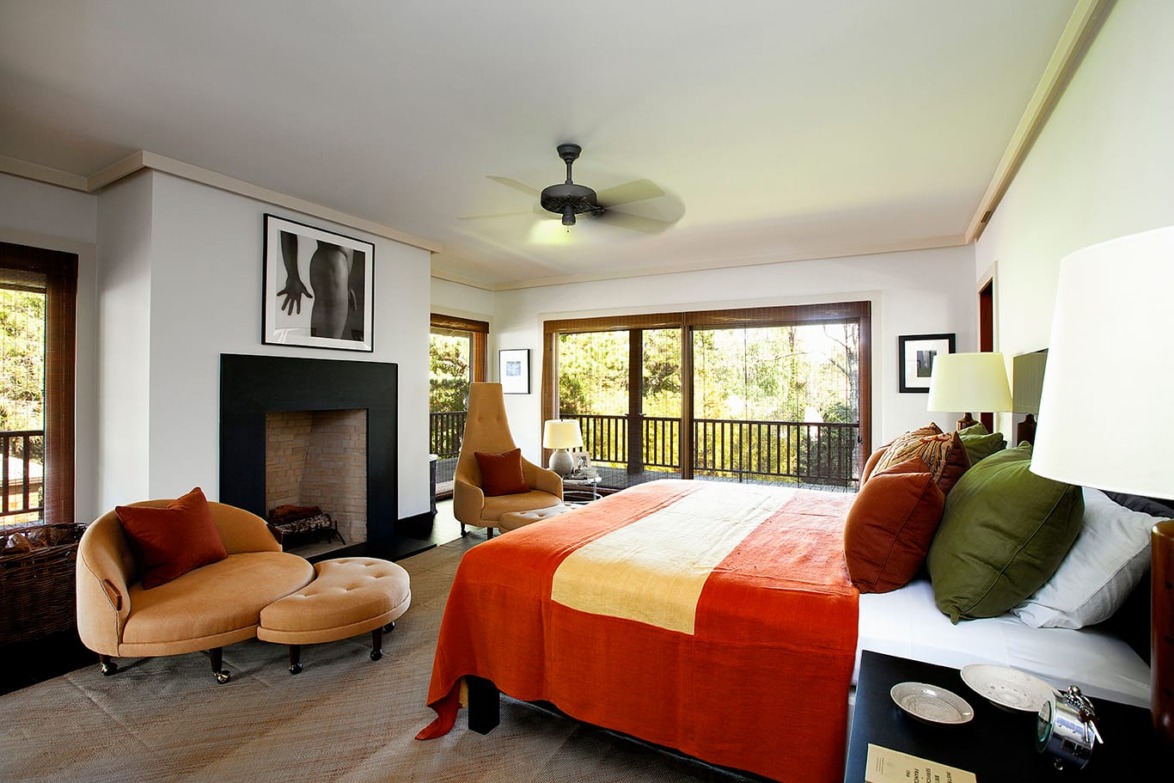 Spacious bedroom with large windows, vibrant orange bedding, cozy seating area, and decorative fireplace.