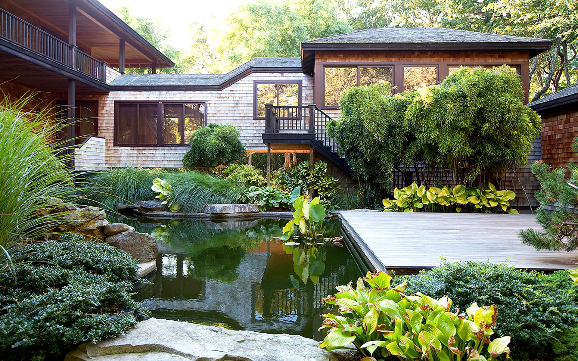 Tranquil backyard pond with surrounding greenery and wooden deck, adjacent to a rustic two-story house.