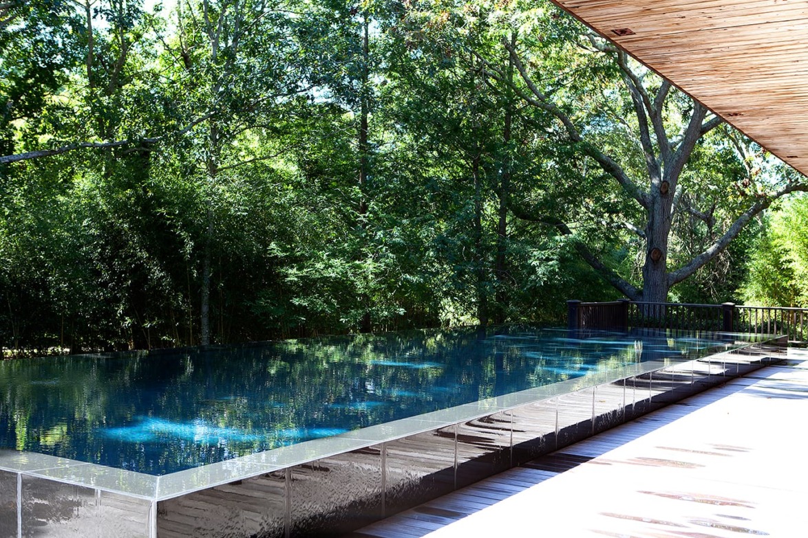 Infinity pool on a wooden deck surrounded by lush green trees under a clear blue sky.