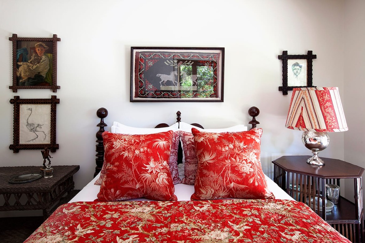 Cozy bedroom with red floral bedding, ornate pillows, framed art on walls, and vintage lamp on dark wood nightstand.