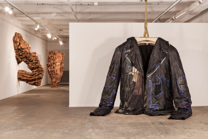 Art exhibit featuring a large black coat sculpture hanging in a gallery with abstract wall sculptures in the background.