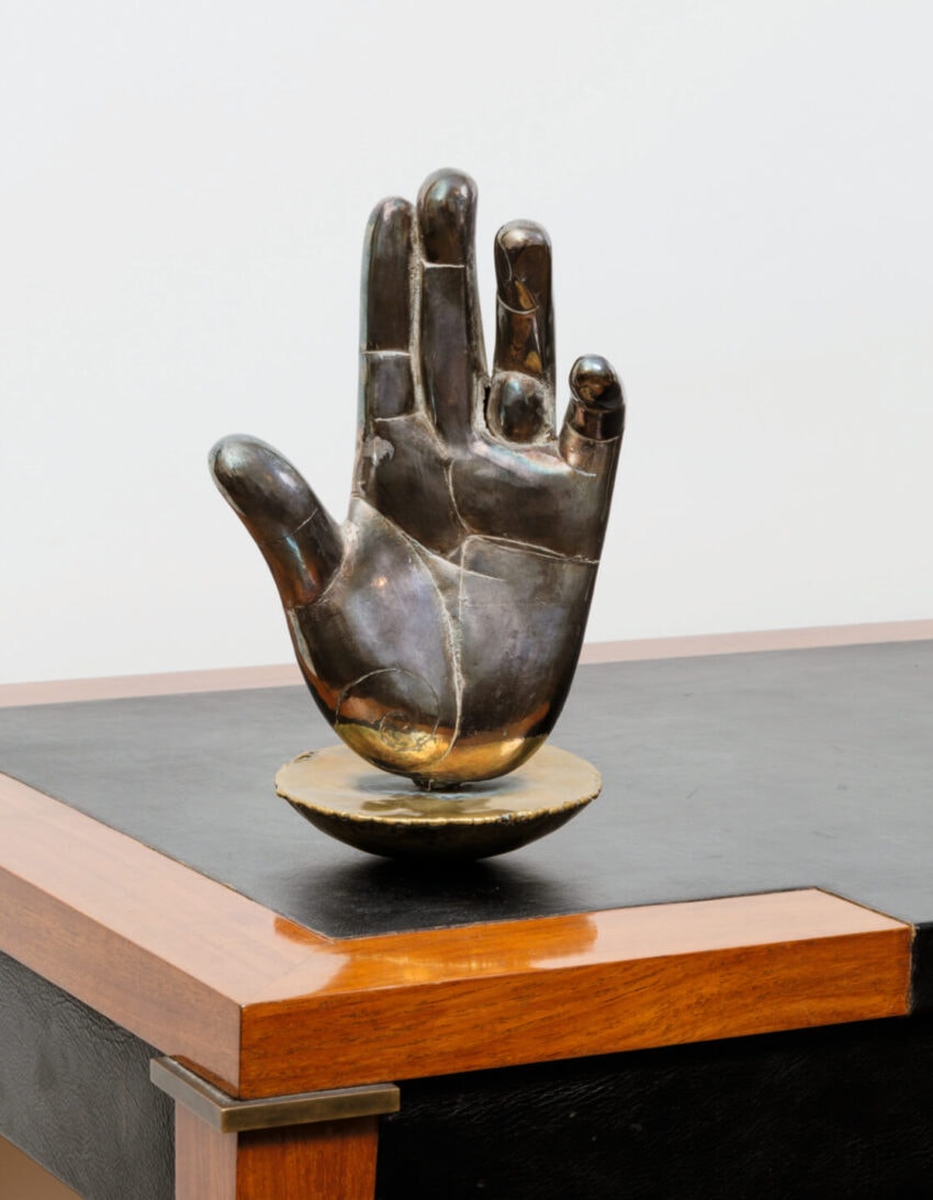 Sculpture of a raised hand with a smooth, reflective surface on a wooden table, resembling a gesture or wave.