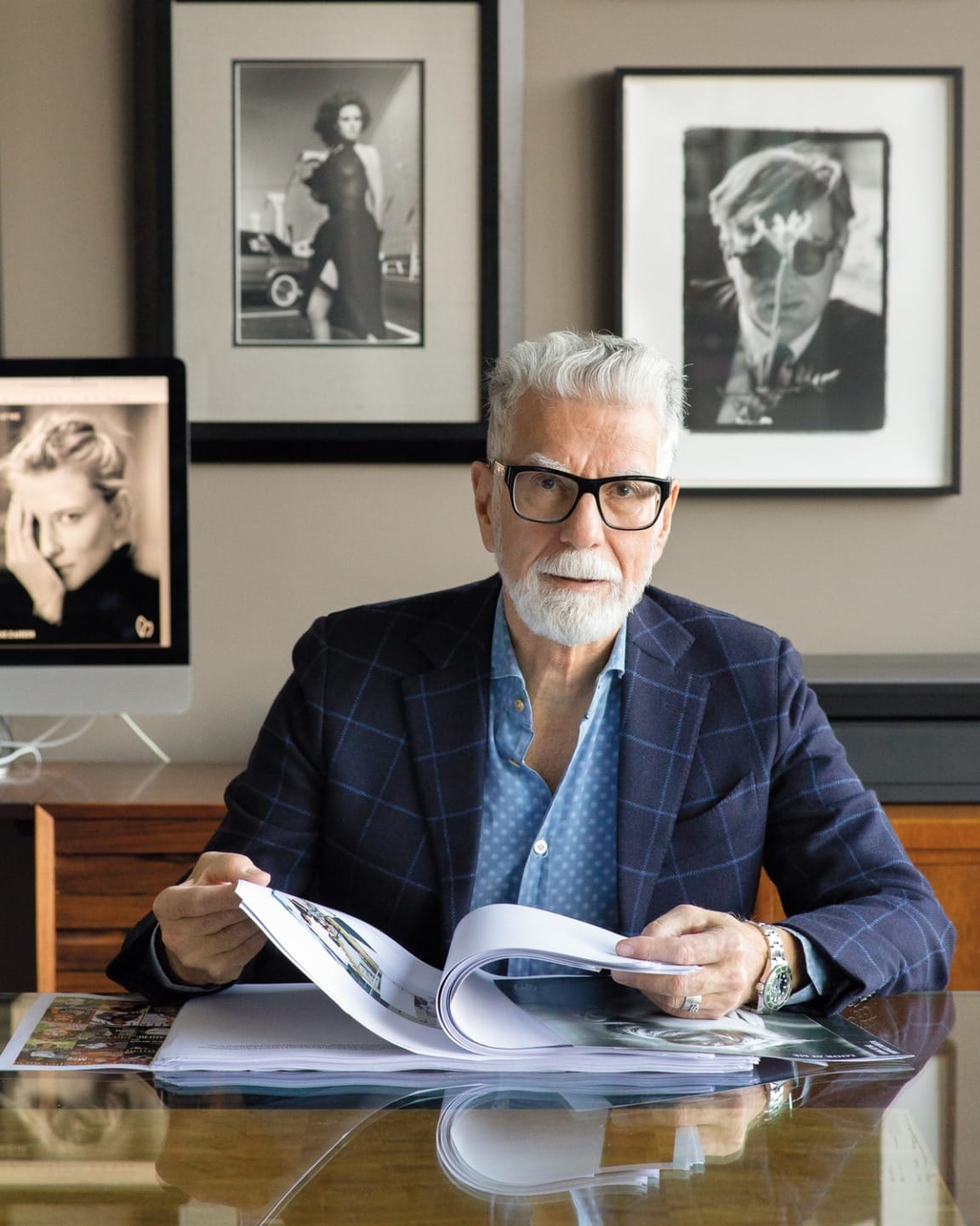 Man with gray hair and glasses seated at a desk with open book, surrounded by framed photos and a computer monitor.